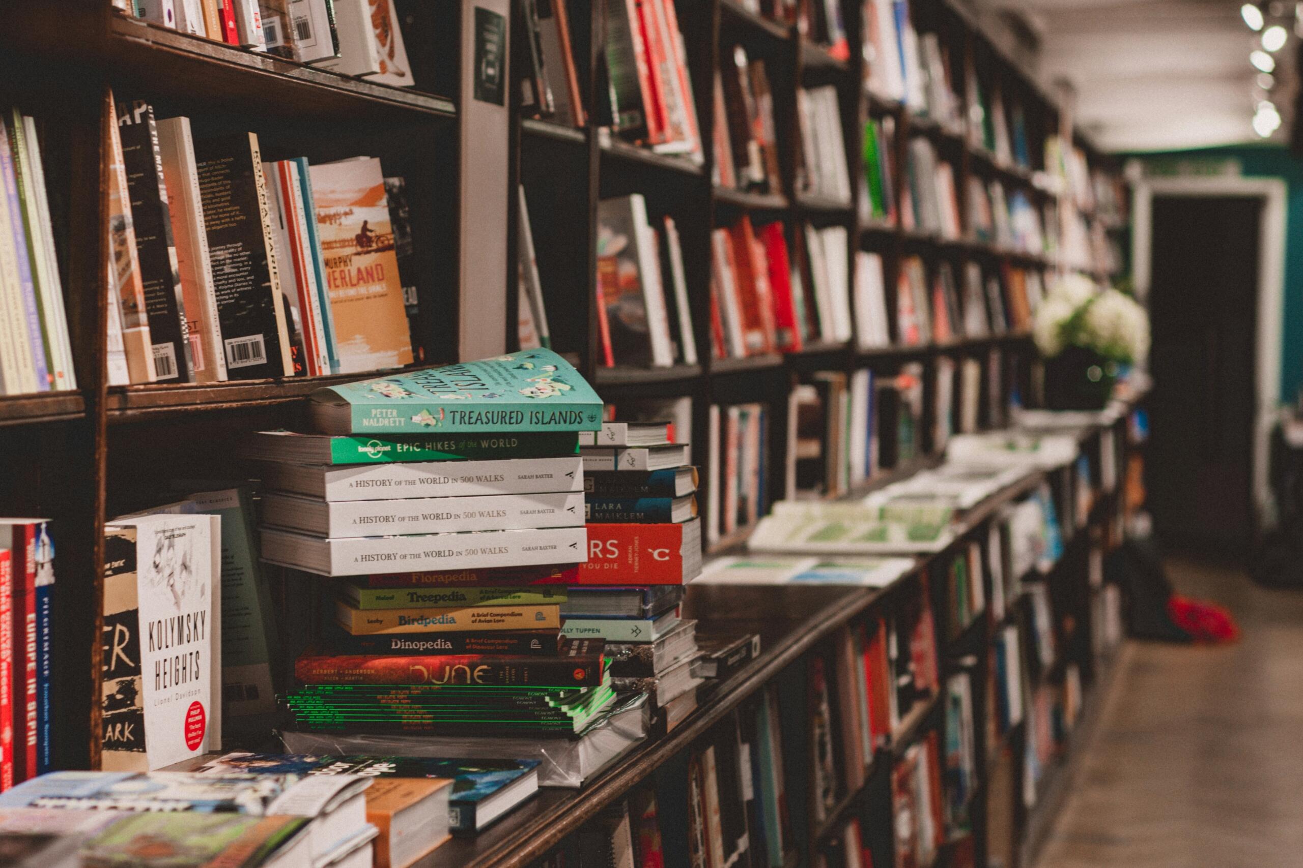 رفوف خشبية طويلة مليئة بالكتب المتنوعة في مكتبة مع كومة من الكتب المكدسة في المقدمة.