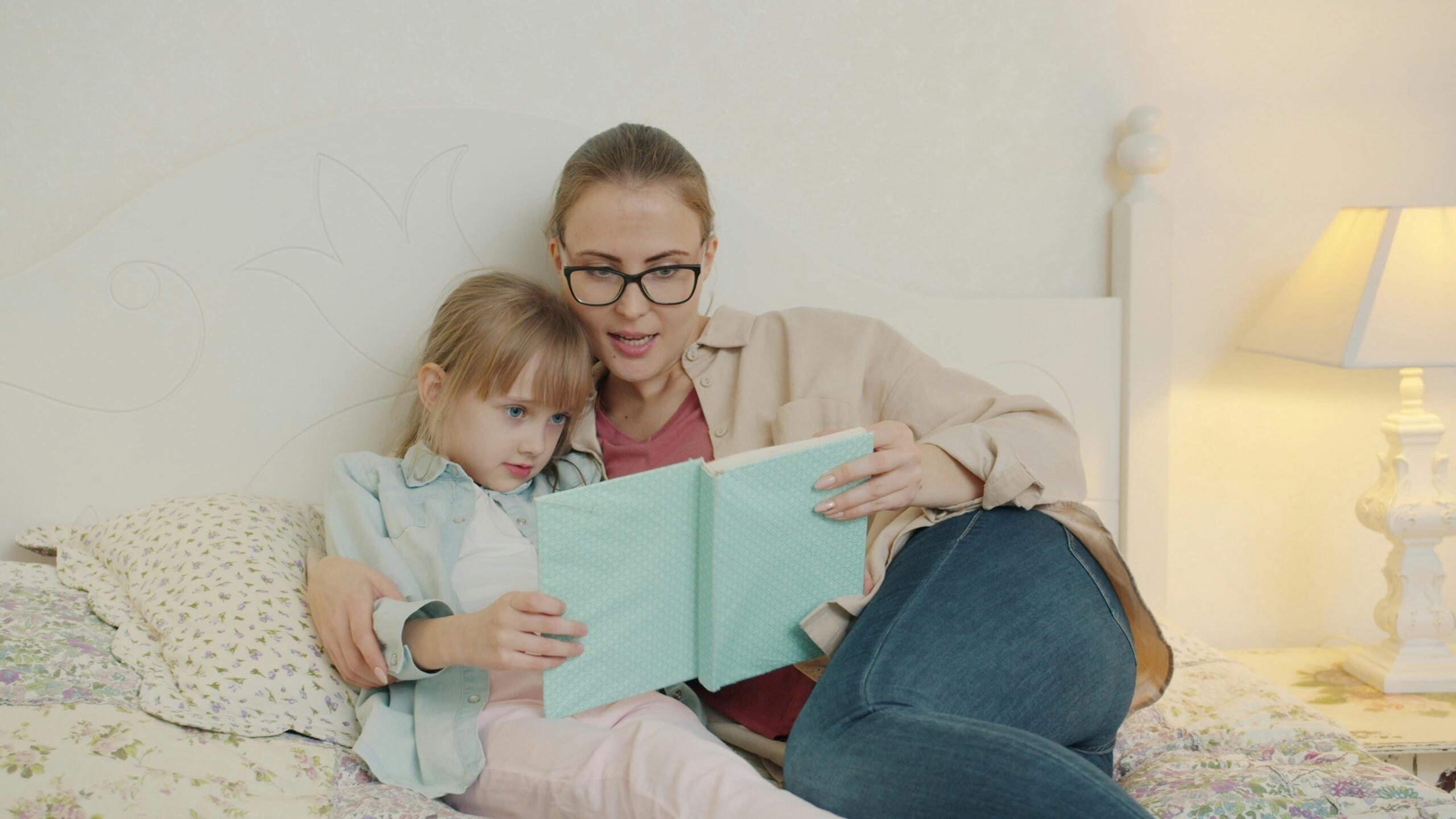 أم تقرأ قصة لطفلتها الصغيرة بتركيز وهدوء أثناء جلوسهما معا على السرير في غرفة النوم.