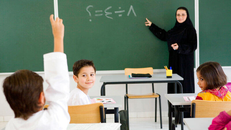 معلمة تشرح على السبورة ، وتتفاعل مع طلابها في الفصل الدراسي بينما يرفع أحد الطلاب يده للمشاركة. يعكس ذلك أهمية تطبيق العلوم المعرفية في التعليم.
