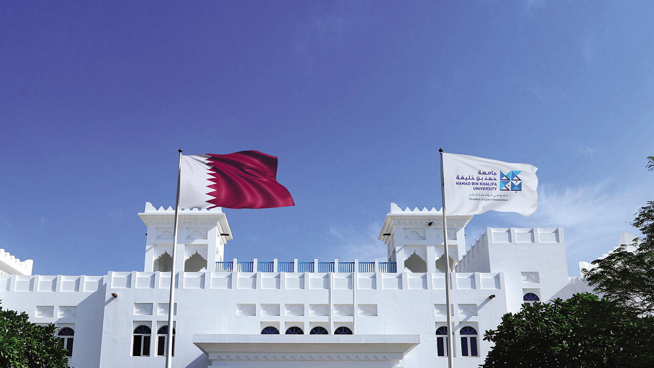 صورة لعلم دولة قطر وعلم جامعة حمد بن خليفة يرفرفان أمام مبنى الجامعة الأبيض ذي العمارة التقليدية.