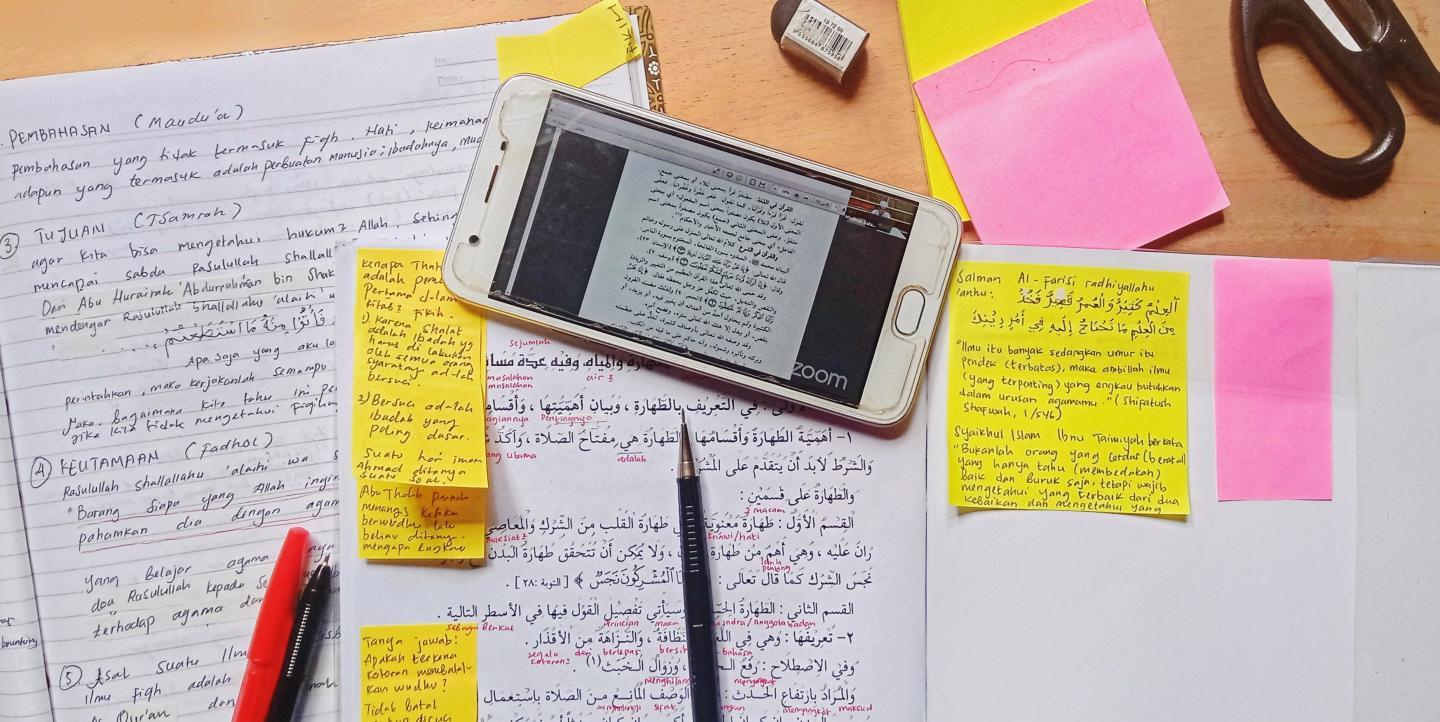 لقطة علوية لمكتب دراسي يعج بالملاحظات والكتب، يظهر فيها كتاب مفتوح إلى جانب دفاتر وهاتف محمول وأوراق ملاحظات ملونة ومكتوبة بخط اليد. مما يجسد عملية تدوين الملاحظات الدراسية وترسيخ المعلومات.