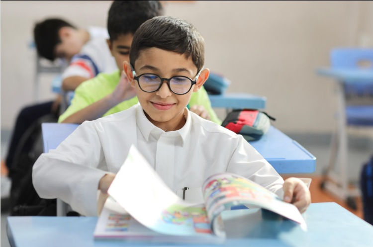 طالب يرتدي نظارة يقلب صفحات كتاب مدرسي بابتسامة وارتياح داخل الفصل، يعبر عن تأقلم الطلبة في المدرسة.