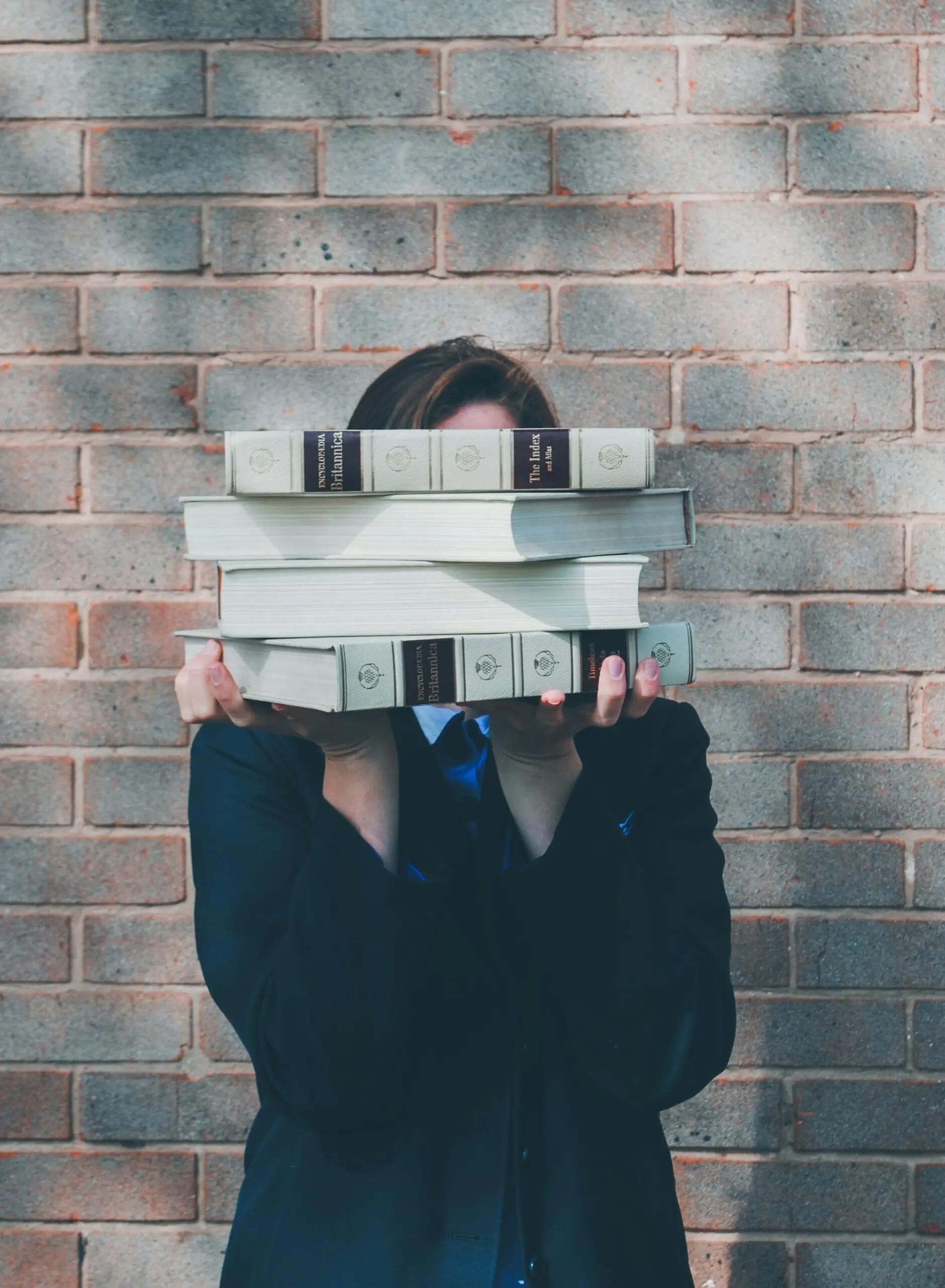 شخص يحمل كومة من الكتب السميكة أمام وجهه وهو يقف أمام جدار مبني من الطوب الأحمر