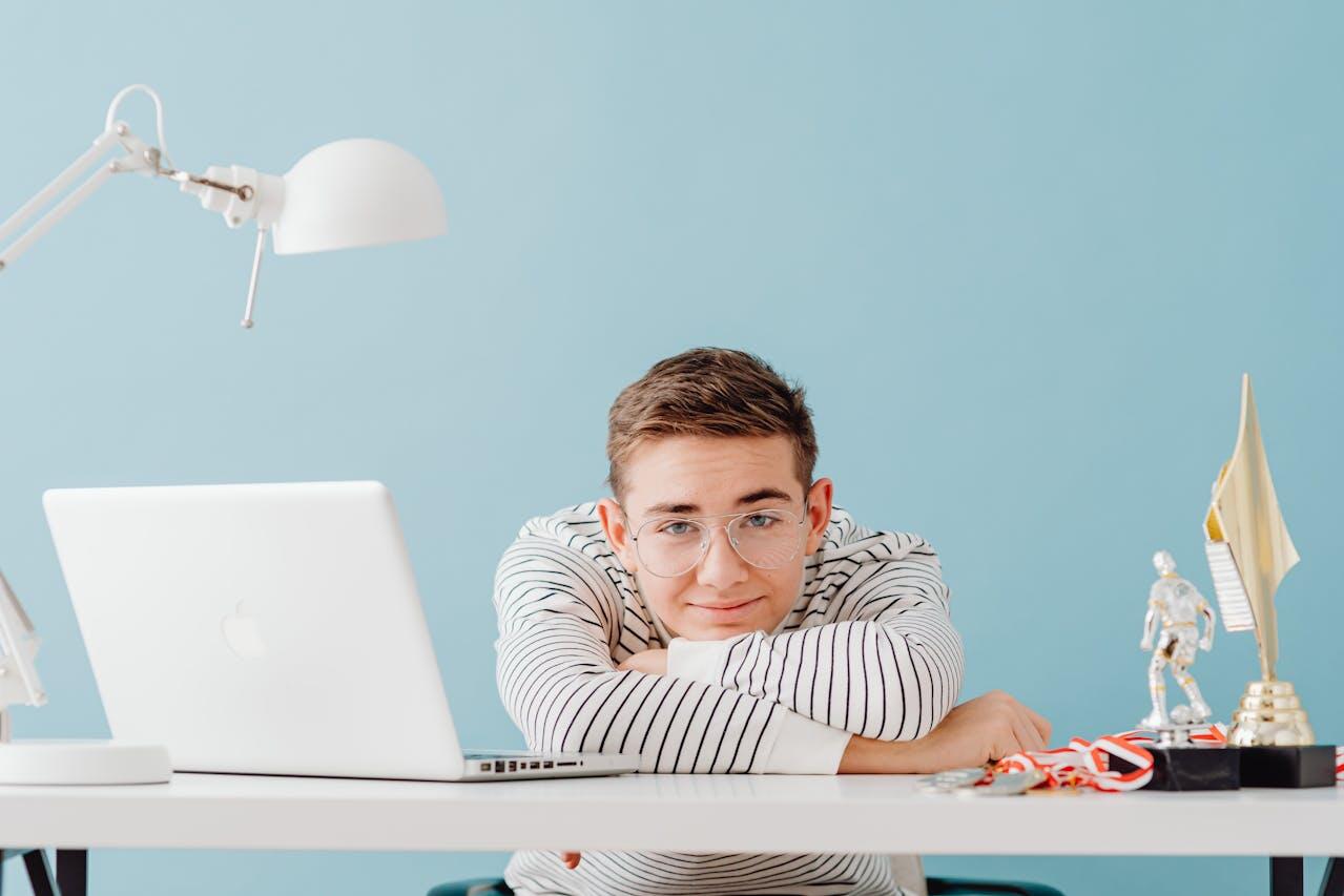 jeune homme a haut raye etale sur son bureau blanc avec lampe et laptop de meme couleur avec un mur bleu ciel en fond