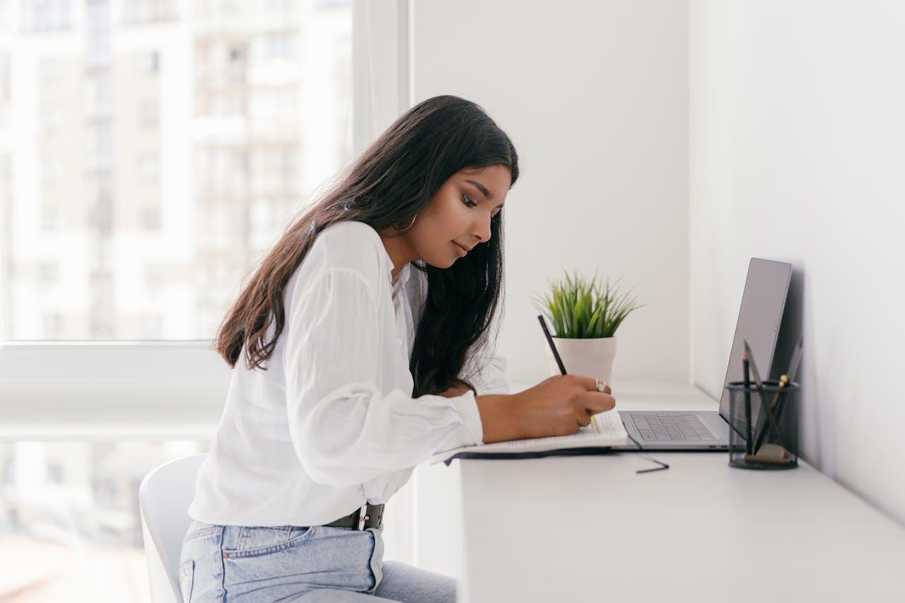 jeune fille aux cheveux long et noir avec une chemise blanche travaillant sur son bureau avec un laptop un crayon et un cahier