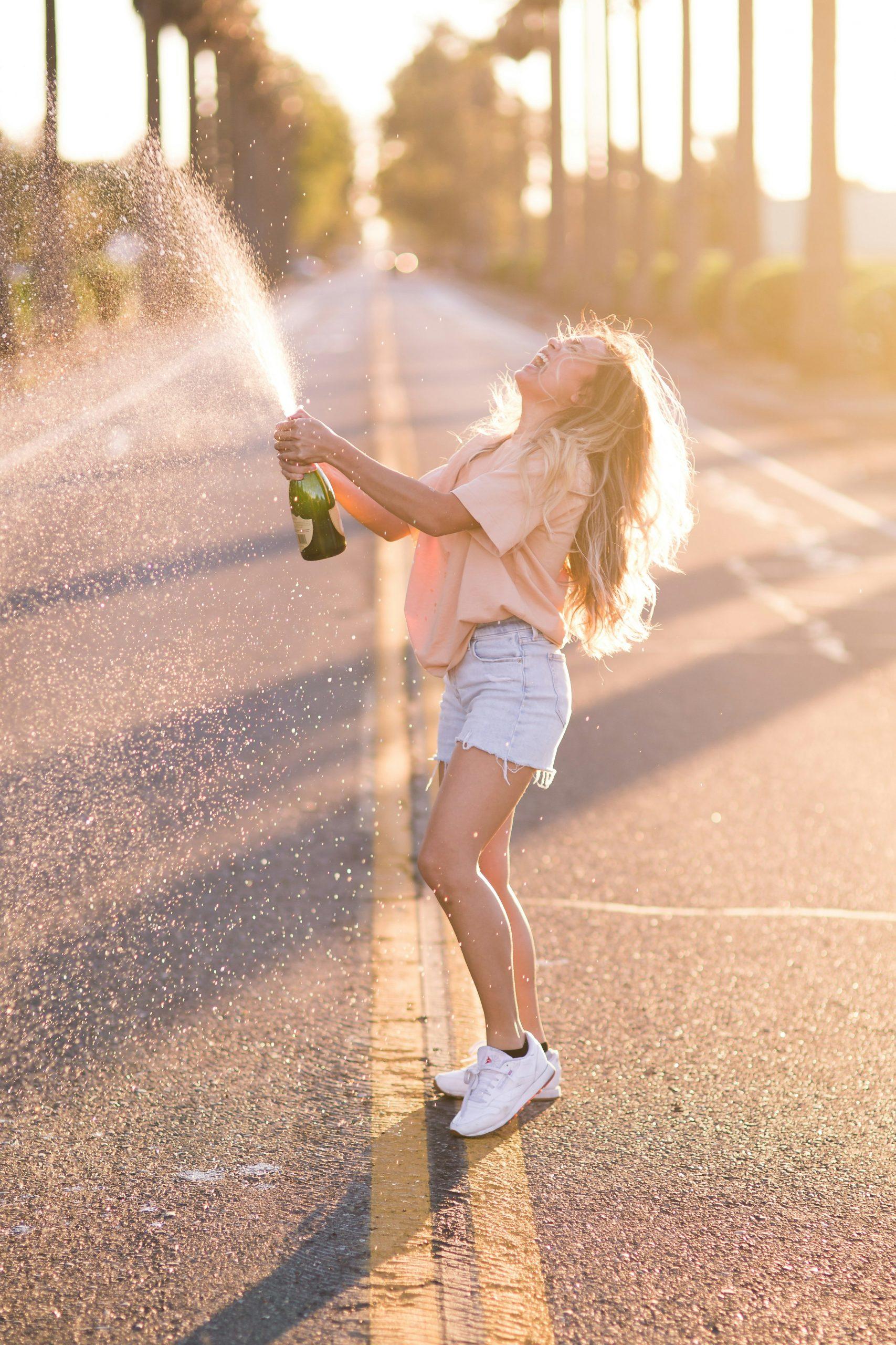 Une jeune femme débouche une bouteille de champagne au milieu d'une route.