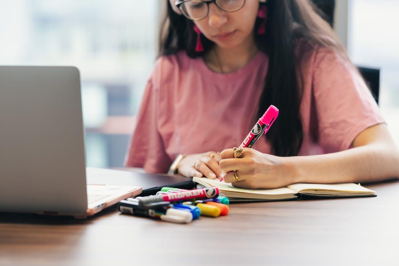 jeune fille aux cheveux noirs et un tee shirt rose qui ecrit sur son cahier avec son laptop ouvert