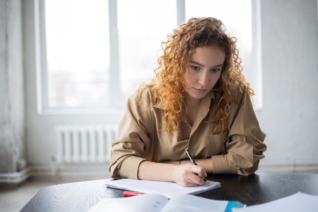 fille rousse aux cheveux boucle concentree en ecrivant sur son cahier sur une table