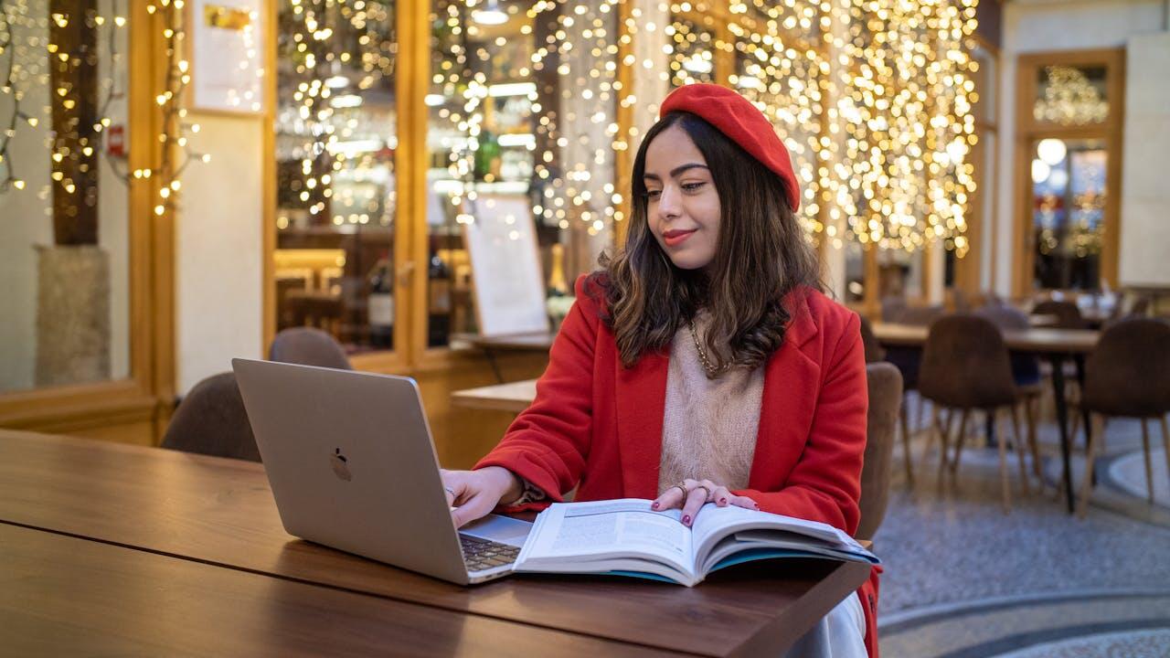 jeune femme a la veste et au beret rouge en train d etudier sur une table avec ordi et livre