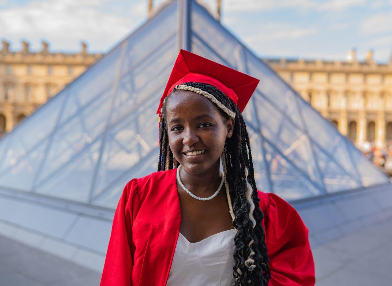 jeune femme de couleur avec toje de diplomee en rouge devant la pyramide du louvre