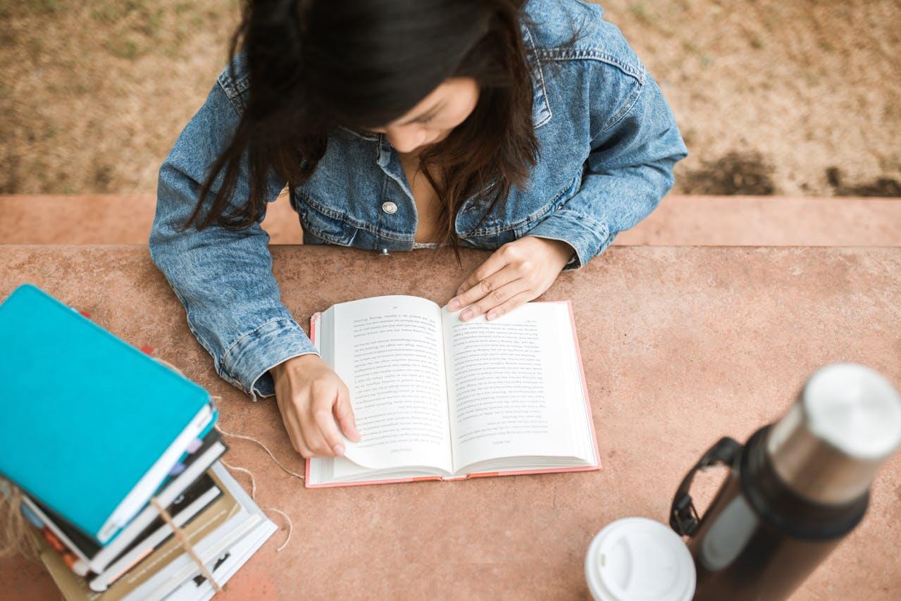 jeune femme en veste en jean a une table lisant avec une pile de livres et une gourde a cote