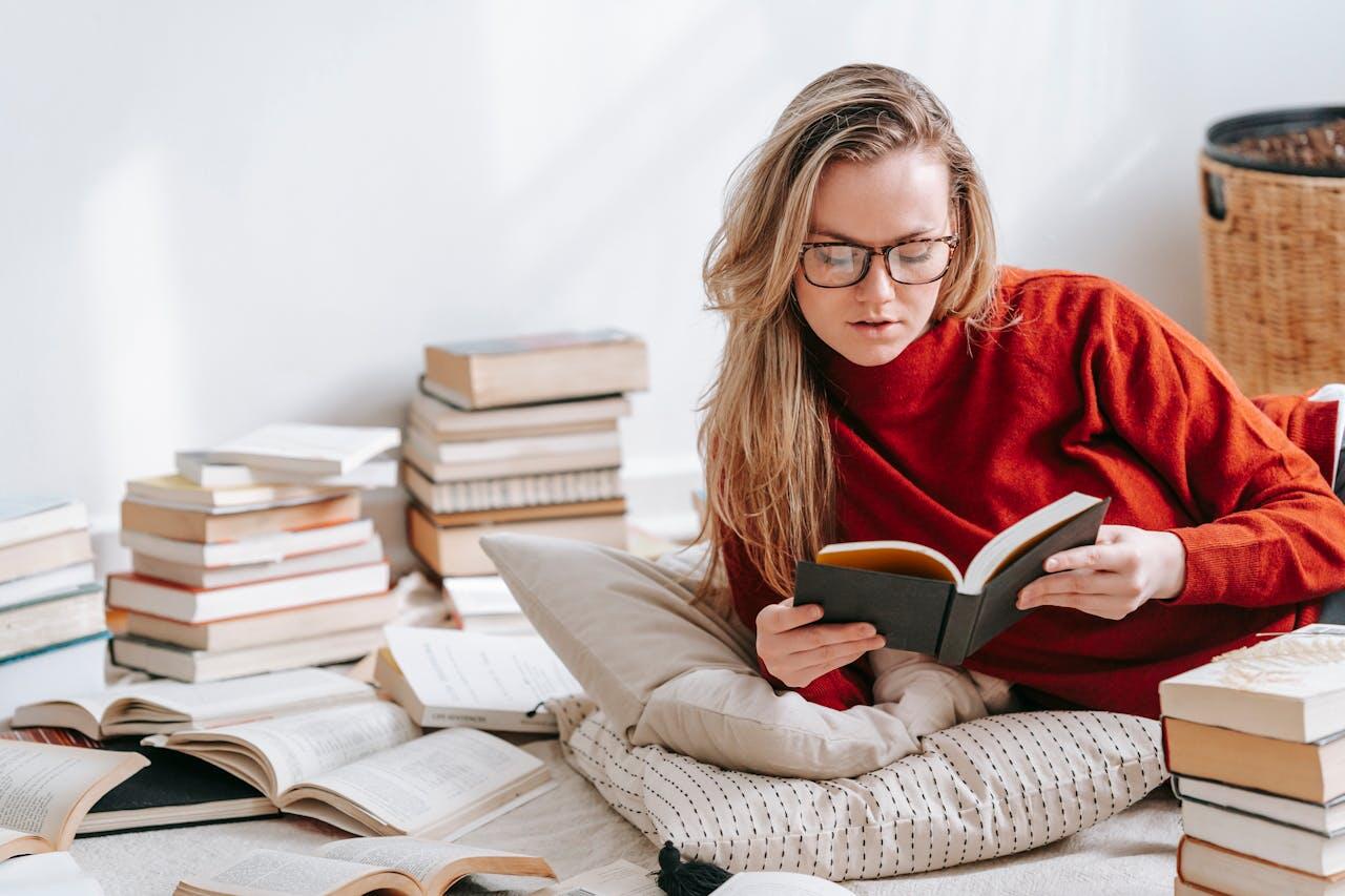 jeune femme blonde a lunette et pull rouge entoures de livres au sol et lisant un livre