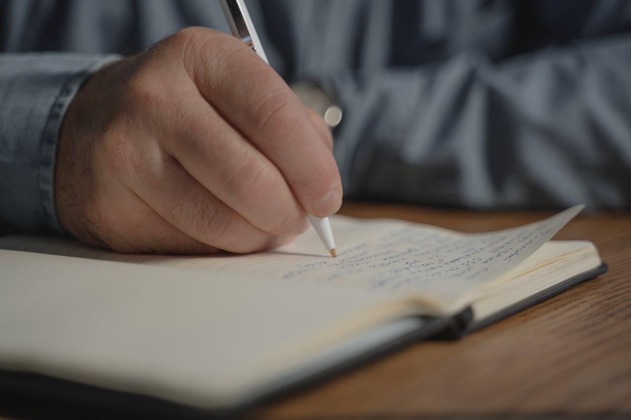 A close-up of a hand holding a pen, writing in a notebook filled with handwritten notes, resting on a wooden table.