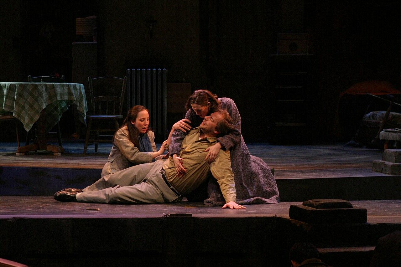 Cast members on stage, one lying on floor, others looking concerned