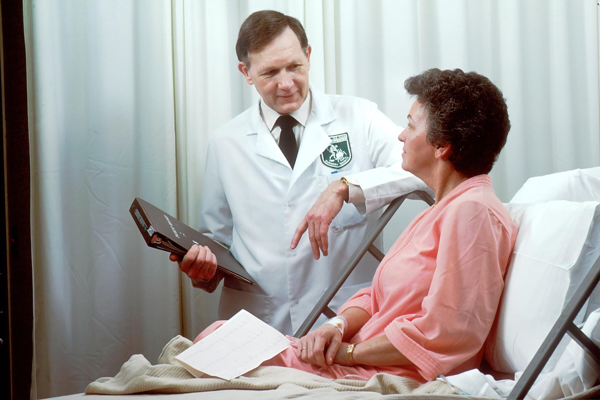 A person in a white lab coat with a green emblem on the left breast stands next to an upraised hospital bed with their elbow propped on its raised rail. They hold a patient file in their right hand while talking with the person in the raised hospital bed, who is wearing a pink robe.