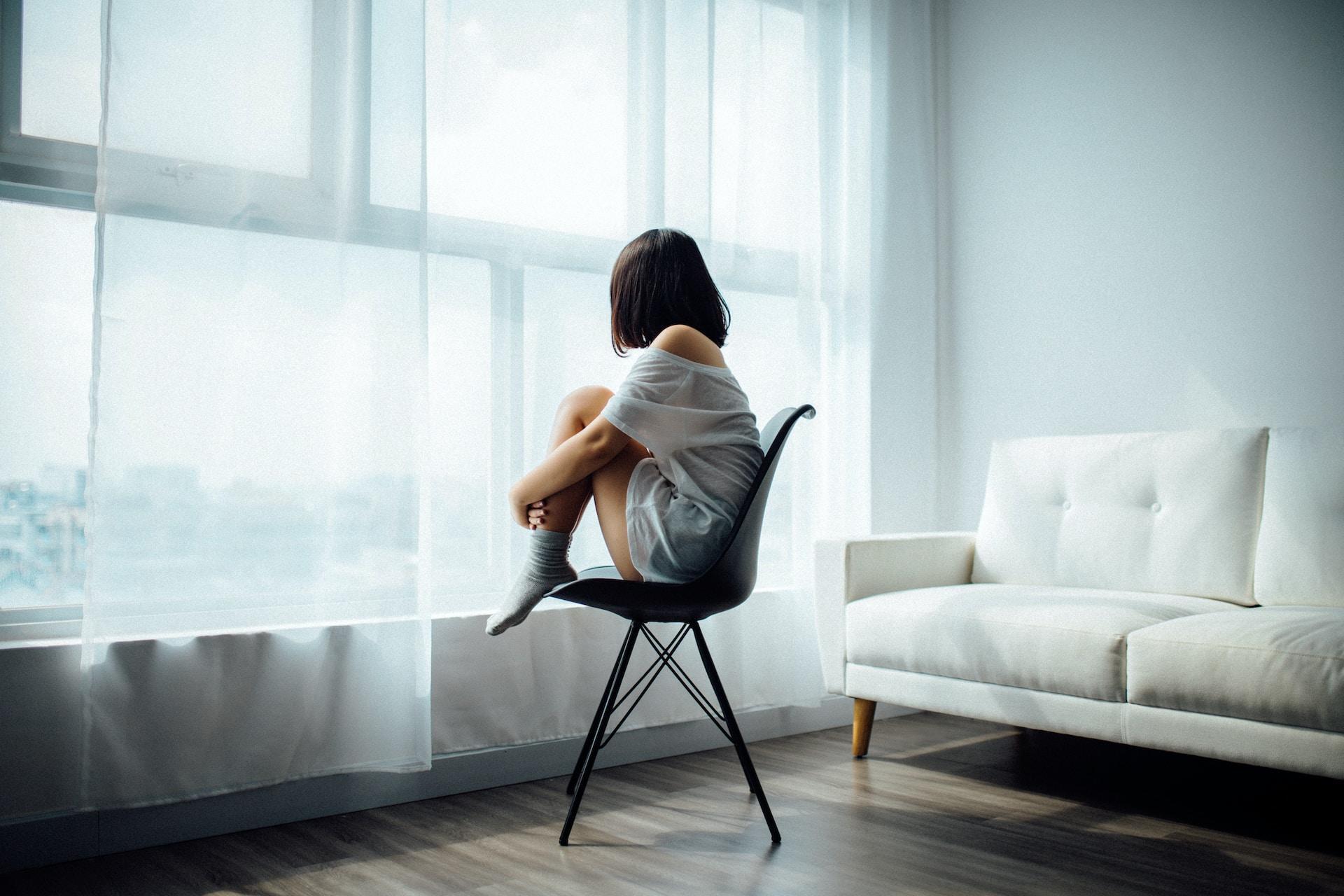 A person wearing a white, gauzy top and grey socks sits on a black chair positioned in front of a white sofa with their knees to their chest, looking out the window.