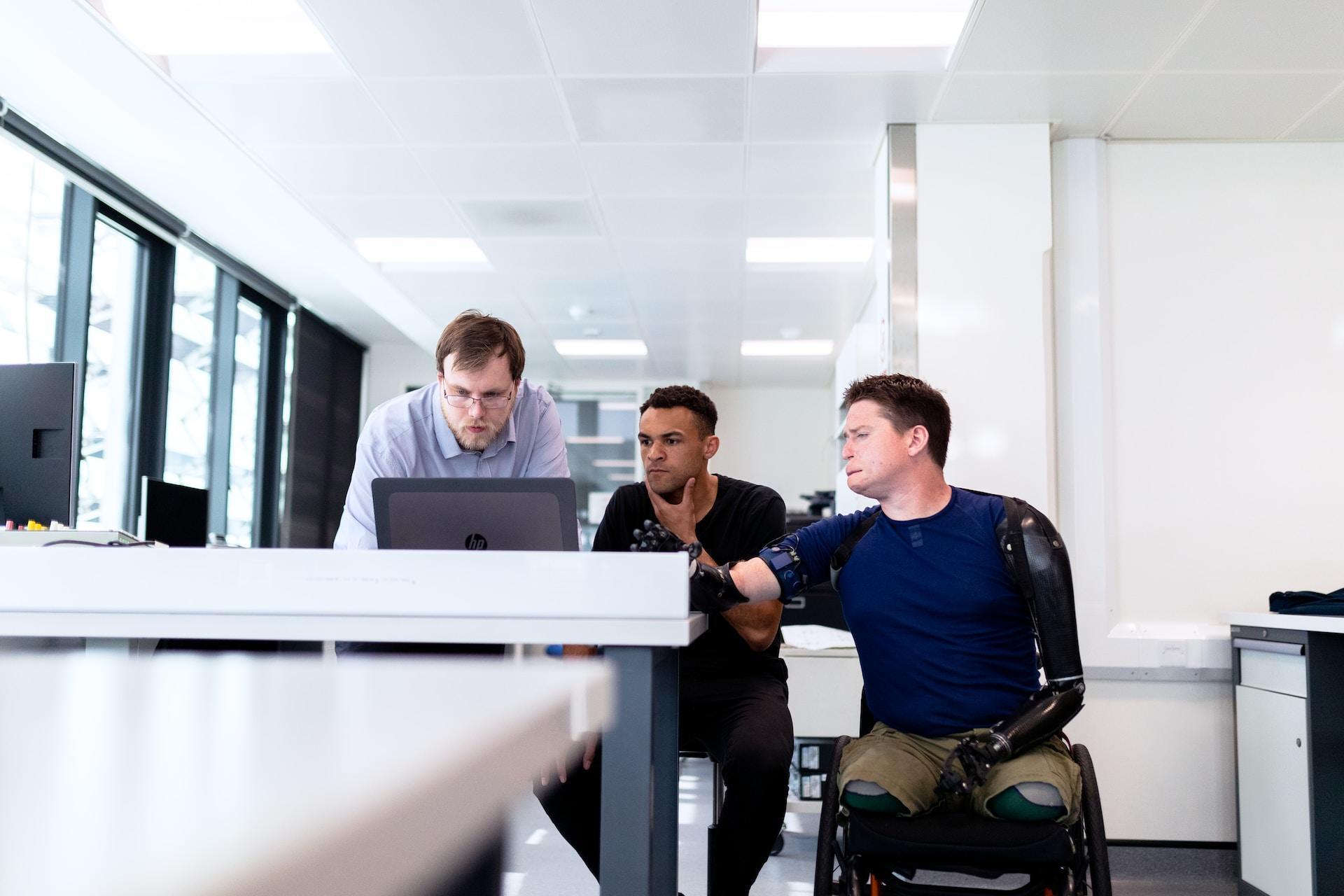 A patient with four robotic appendages sits in a wheelchair and looks at a a computer screen with two other people. He wears a dark blue short sleeved shirt and brown trousers, a sharp contrast in the all-white room.