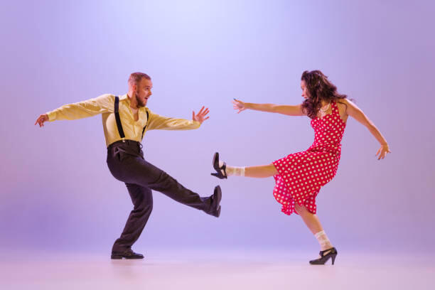 Fată frumoasă și bărbat în costume colorate în stil retro, dansând dansuri incendiare izolate pe fundal liliac în lumină neon. Actori în mișcare și acțiune. Concept de artă, cultura anilor '60, '70.