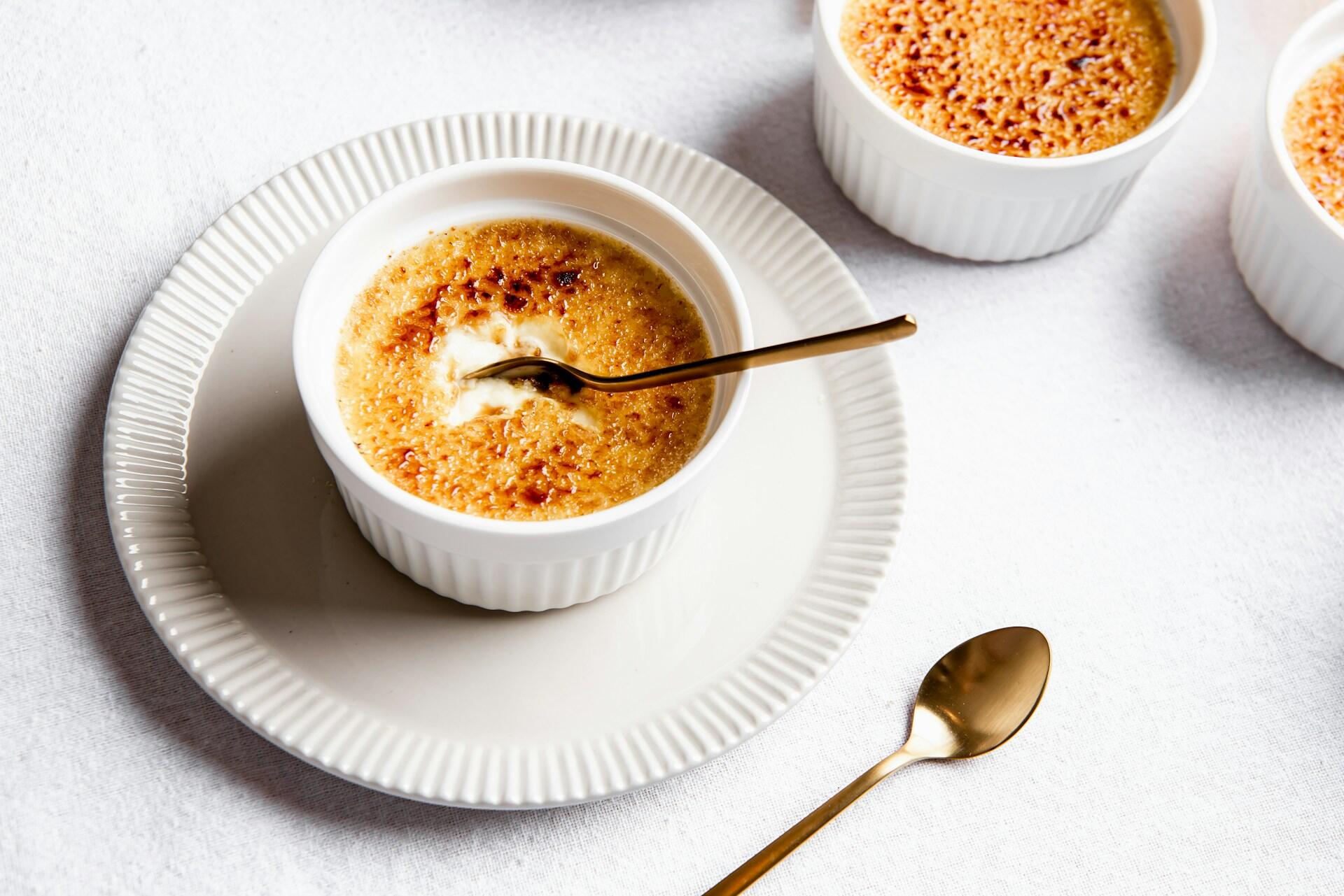 Ett vitt bord med vitt porslin med Crème Brûlée i. I bilden syns även två skedar i guld.