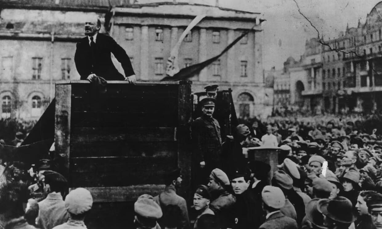 Historisk svartvit bild av en folkmassa samlad kring Lenin på en upphöjd plattform, med byggnader i bakgrunden.