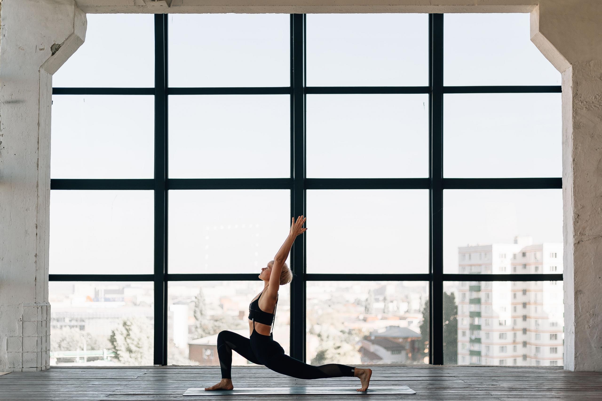 Kvinna i svarta träningskläder gör yoga framför ett stort fönster med storstadsutsikt