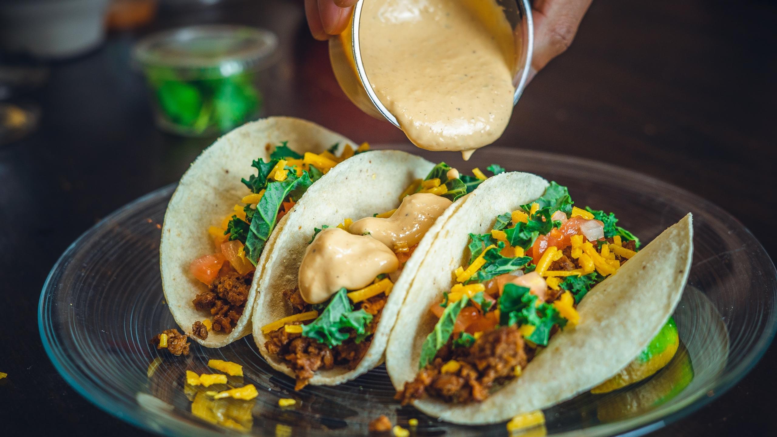 en person häller sås på tre tacos som är fyllda med grönsaker och toppats med koriander