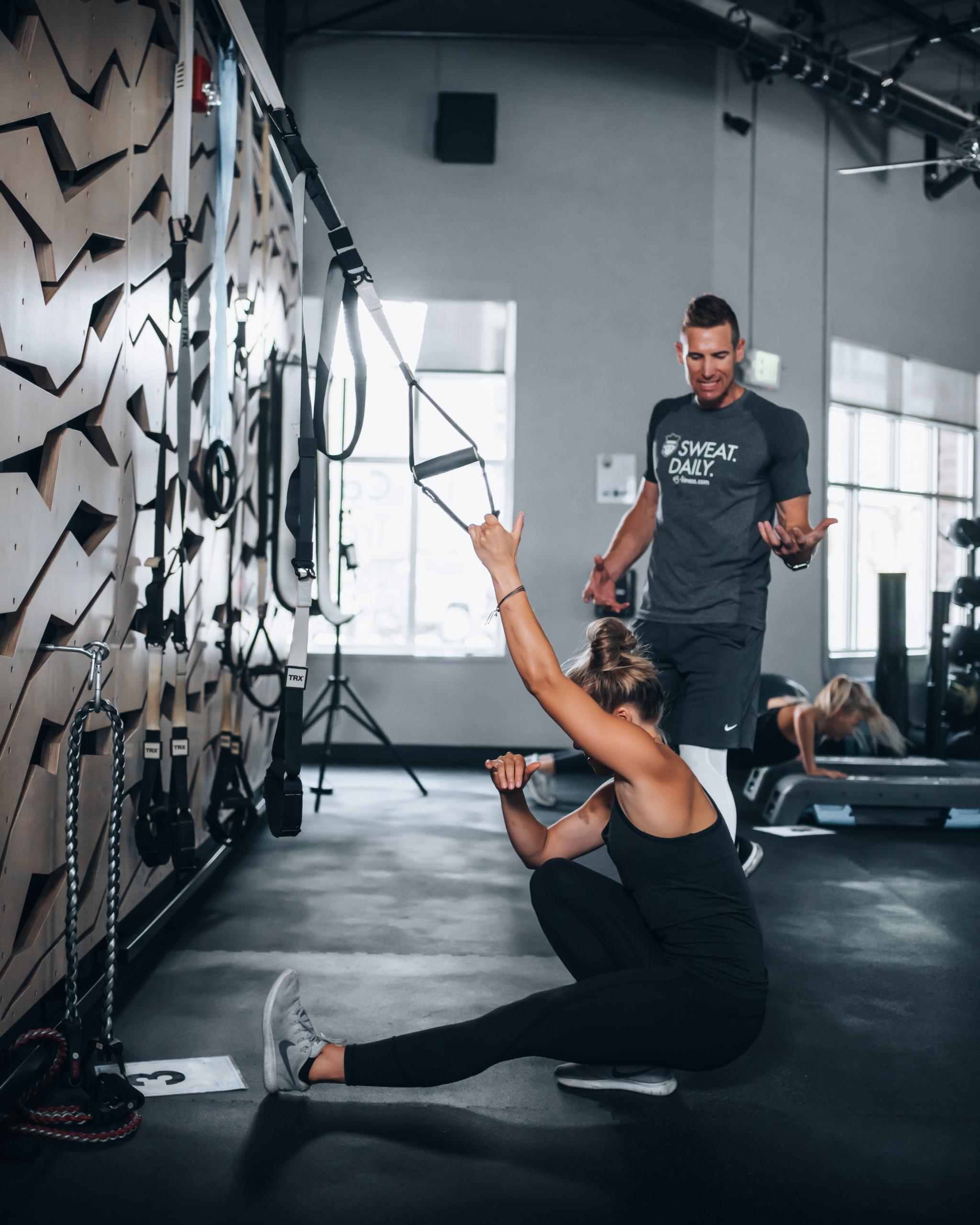 Kvinna gör pistol squat med hjälp av TRX band medan personlig tränare instruerar henne