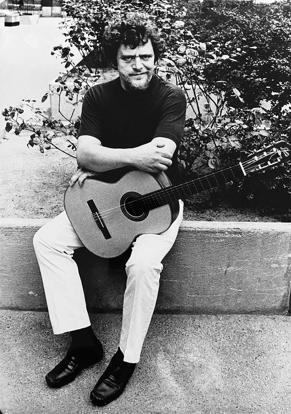 Fred Åkerström sitter på en mur med en gitarr i knät och tittar rakt in i kameran. Bilden är svartvit och i bakgrunden syns en rosenbuske.