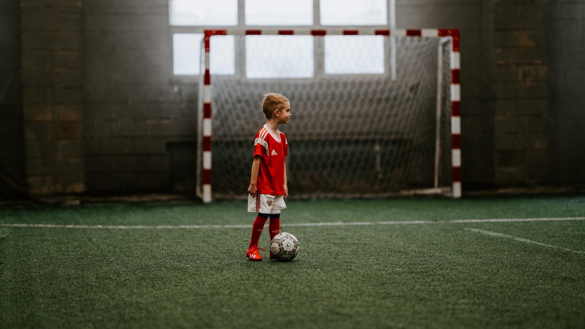 En pojke står på en fotbollsplan inomhus med en boll vid fötterna.