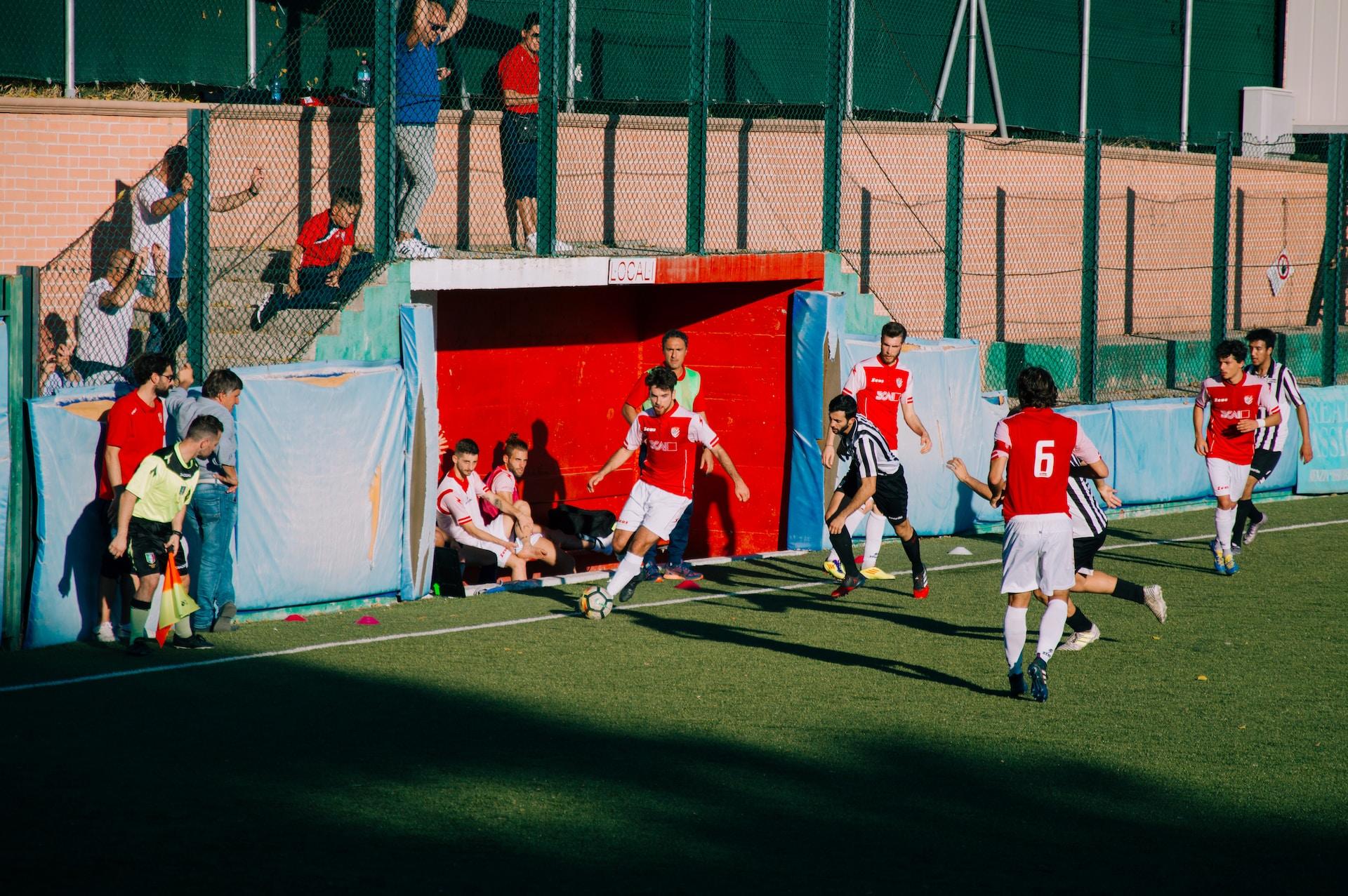 Unga män spelar fotboll på en fotbollsplan med liten läktare. Några står och tittar på.