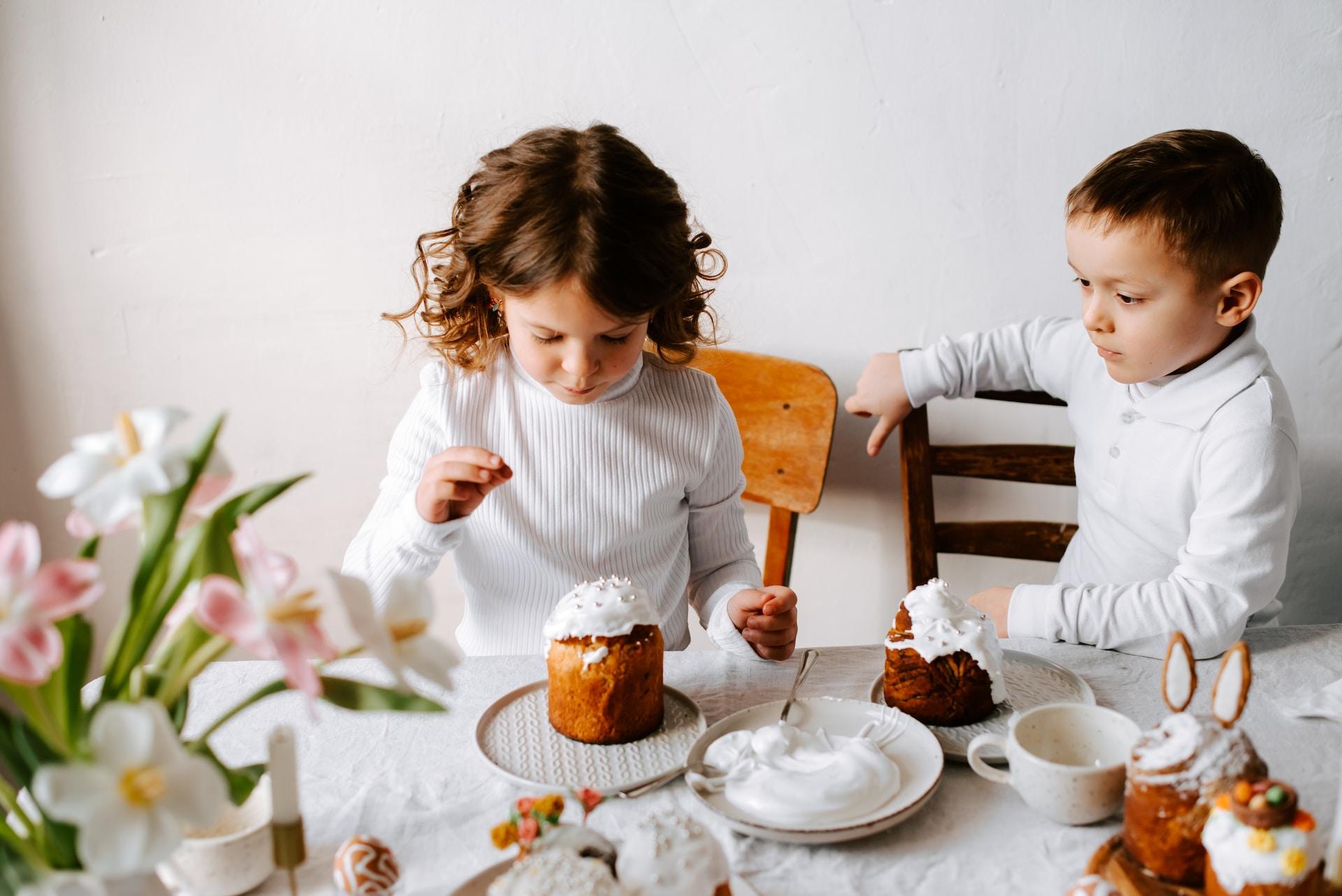 Två barn som sitter vid ett dukat bord. På bordet står bland annat blommor, porslin och kakor.