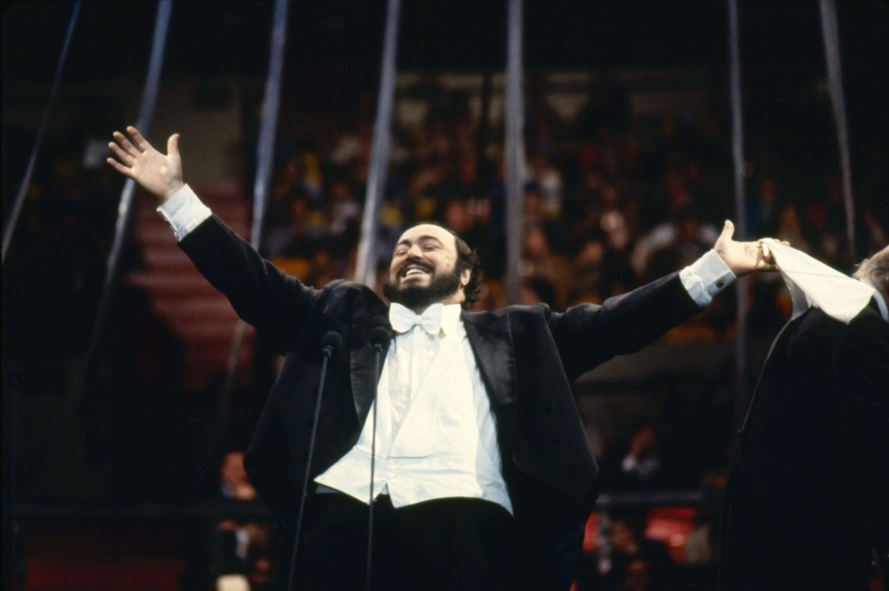Operasångare Luciano Pavarotti står på scenen med utsträckta armar och ett stort leende.