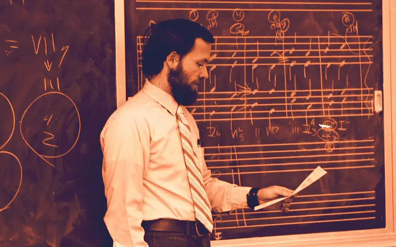 Musiklärare framför en svart krittavla med noter i vit text. 