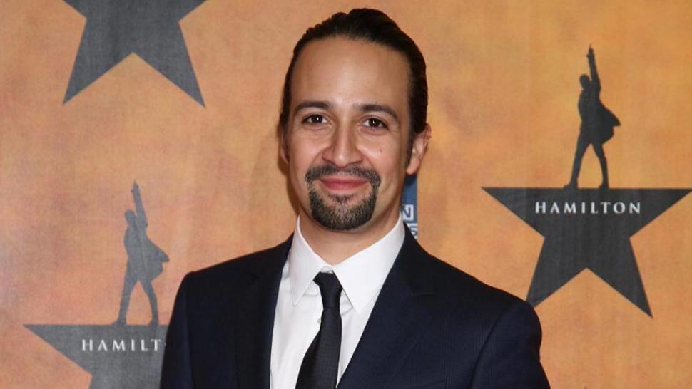 Lin Manuel Miranda i kostym med en Hamilton affisch som bakgrund. 