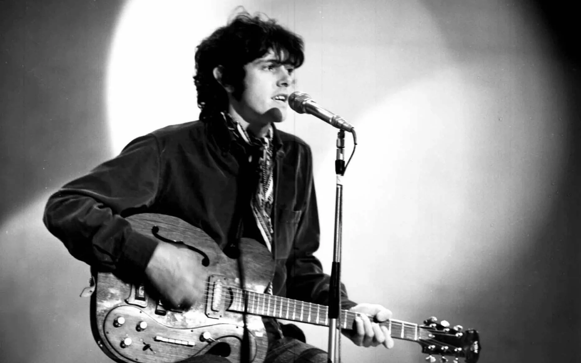 Artisten Donovan sitter på scen med en gitarr i knät.