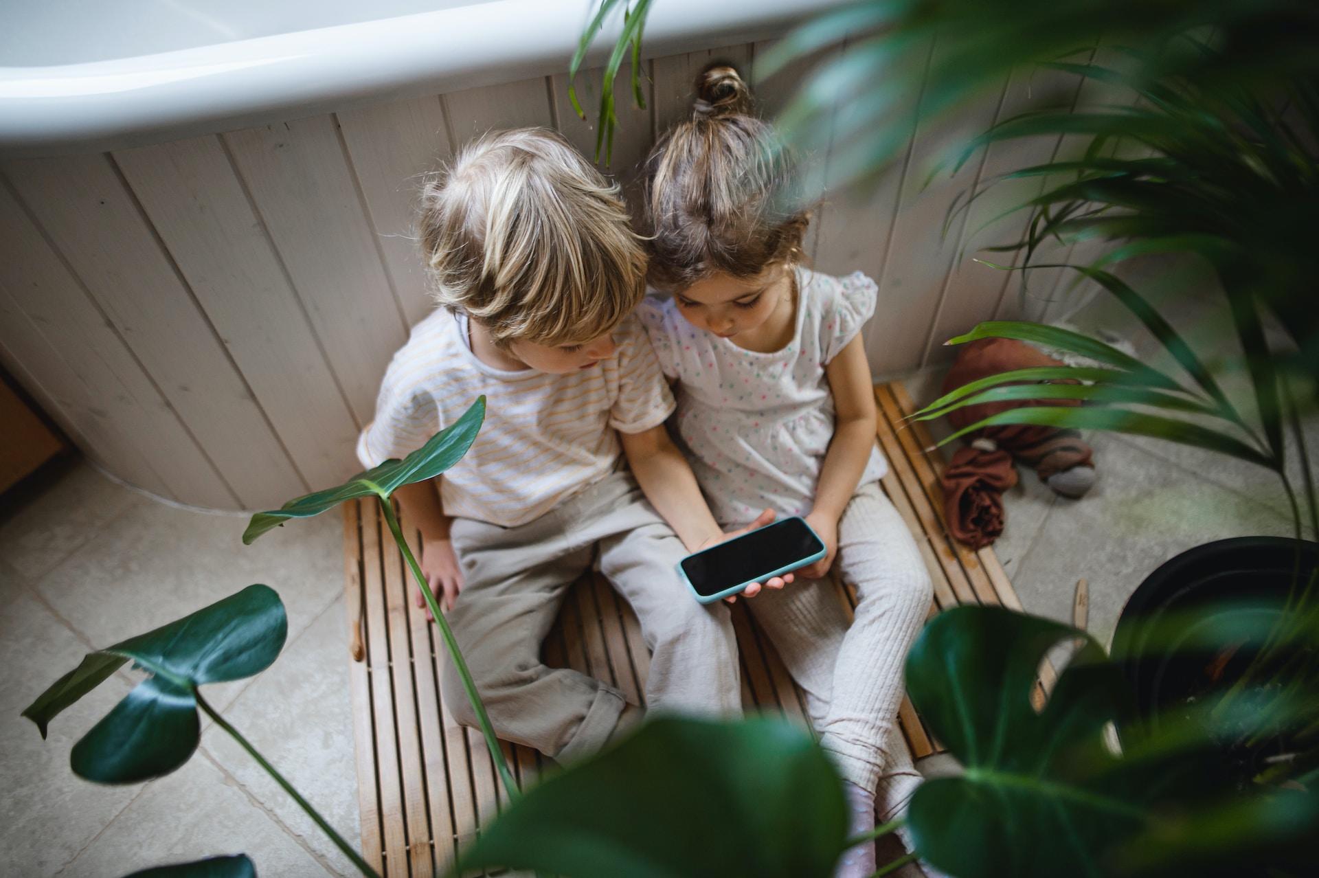Två barn sitter nedanför ett fönster och tittar i en telefon.