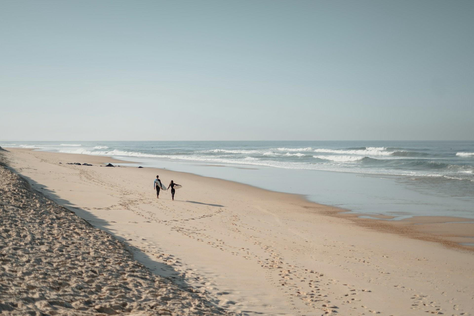 Två surfare som promenerar på stranden.