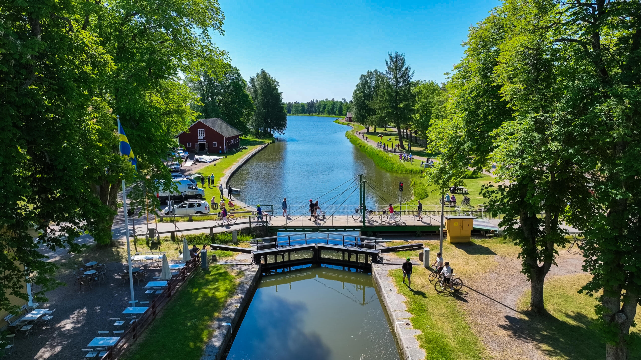 Grönskande träd som växer längst med Göta Kanal. I mitten av bilden syns en sluss och en bro med gående och cyklister.