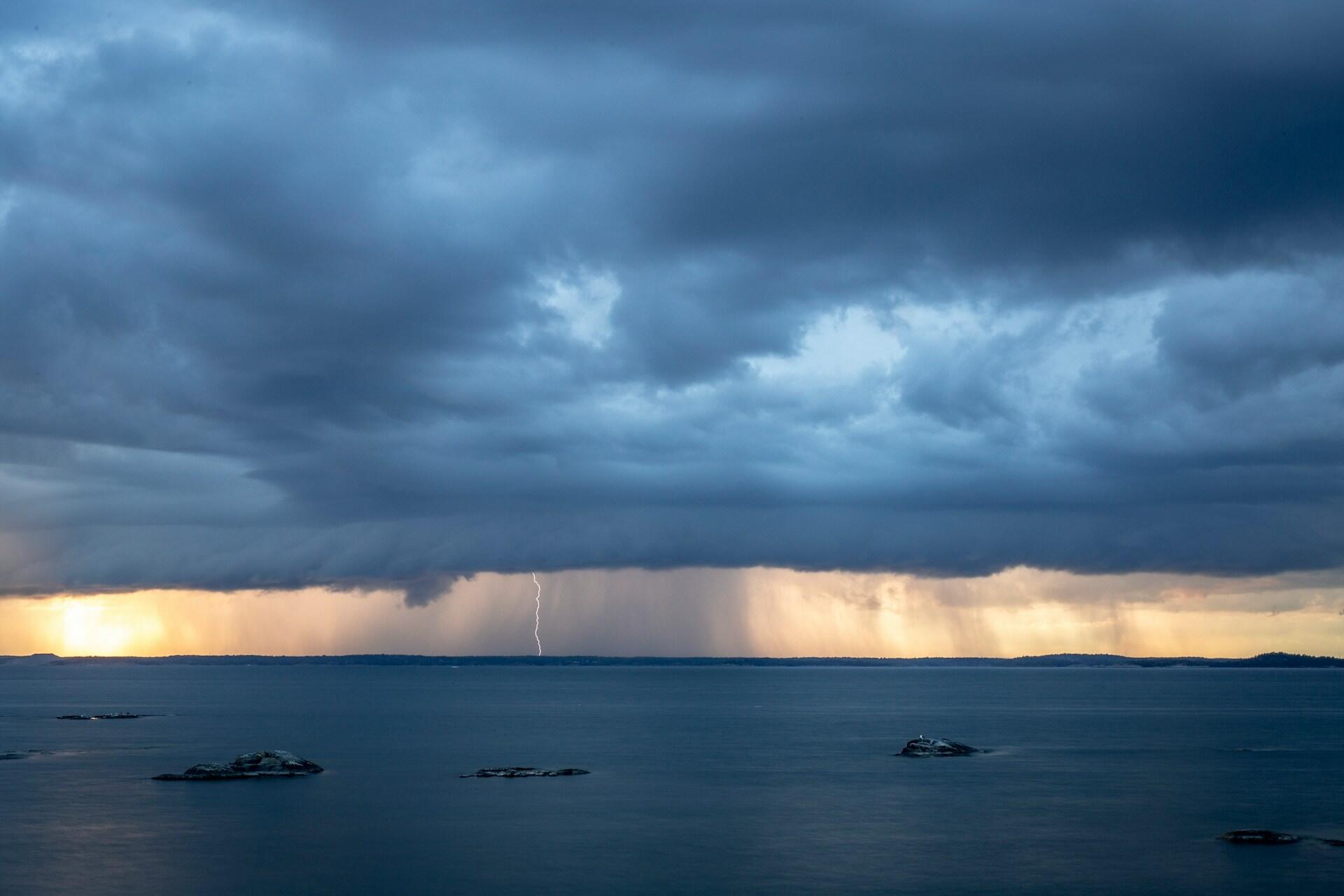 Dramatisk himmel med mörka stormmoln över ett lugnt hav, upplyst av avlägsna blixtar och mjuka solnedgångsfärger vid horisonten.