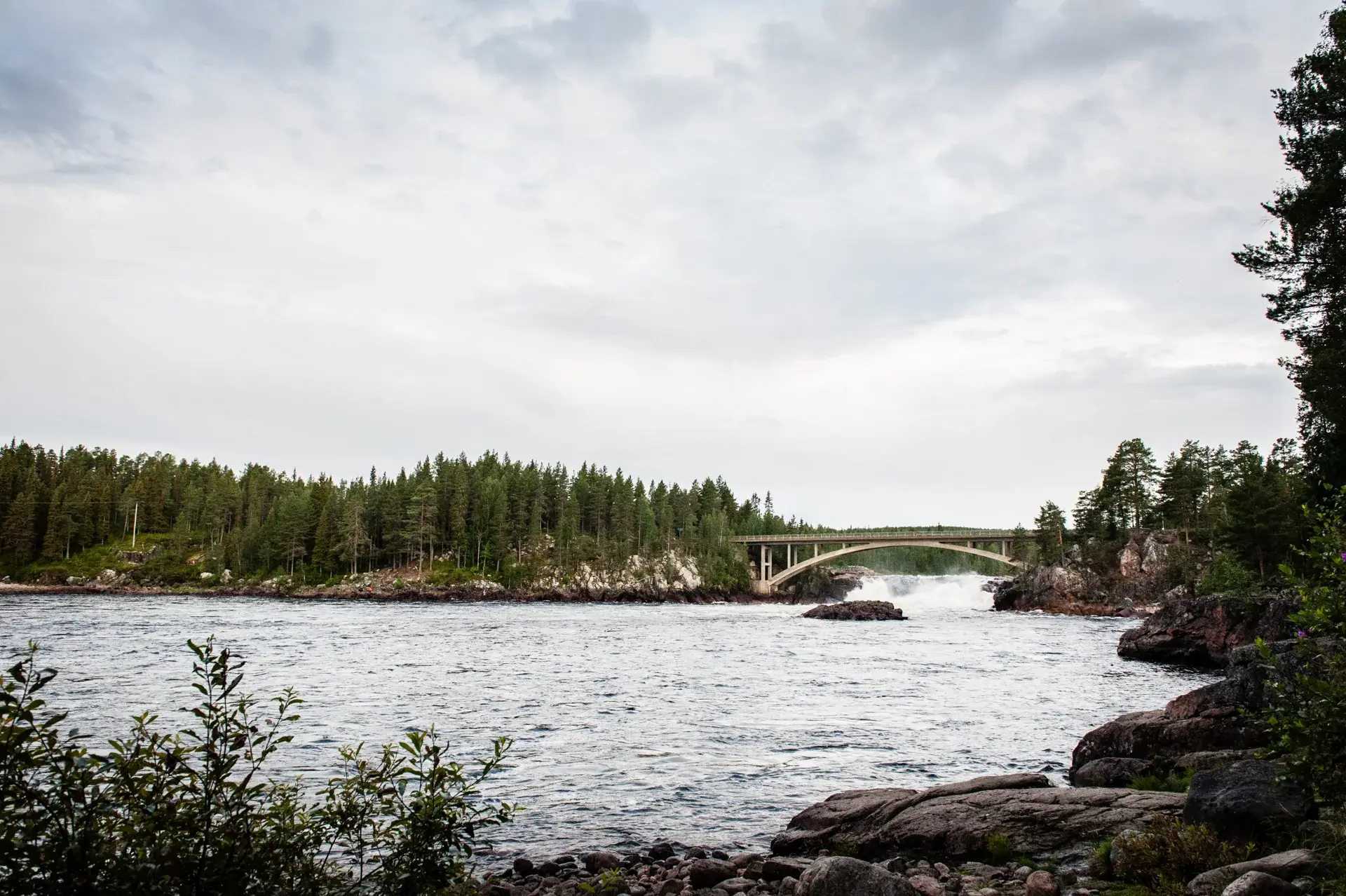 En lugn flod flyter under en böjd bro, omgiven av tät skog och klippiga stränder under ett molnigt himmel.