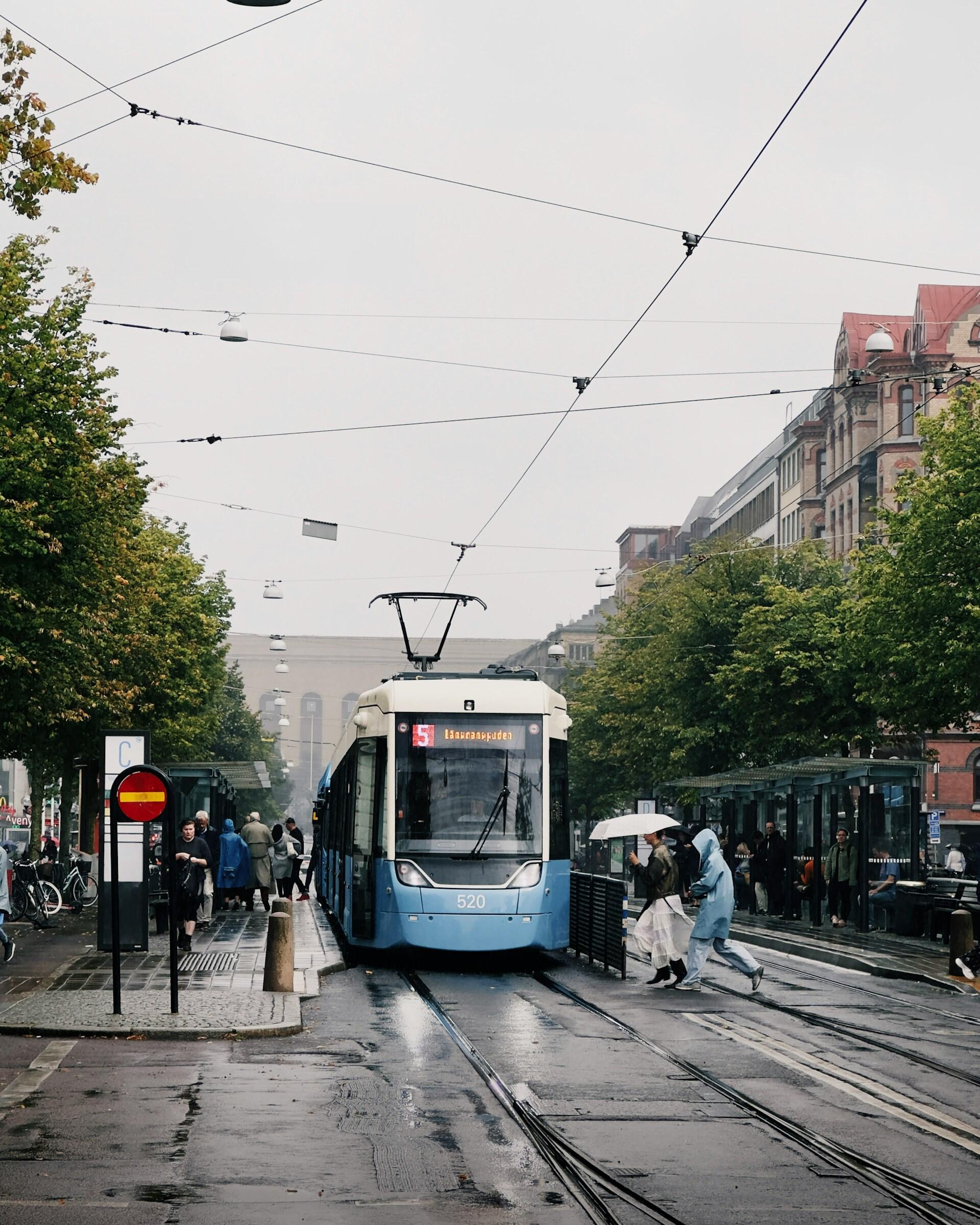 En blå spårvagn stannar vid en regnblöt stadsgata, medan passagerare i regnrockar delar på ett paraply och andra väntar vid en busshållplats.