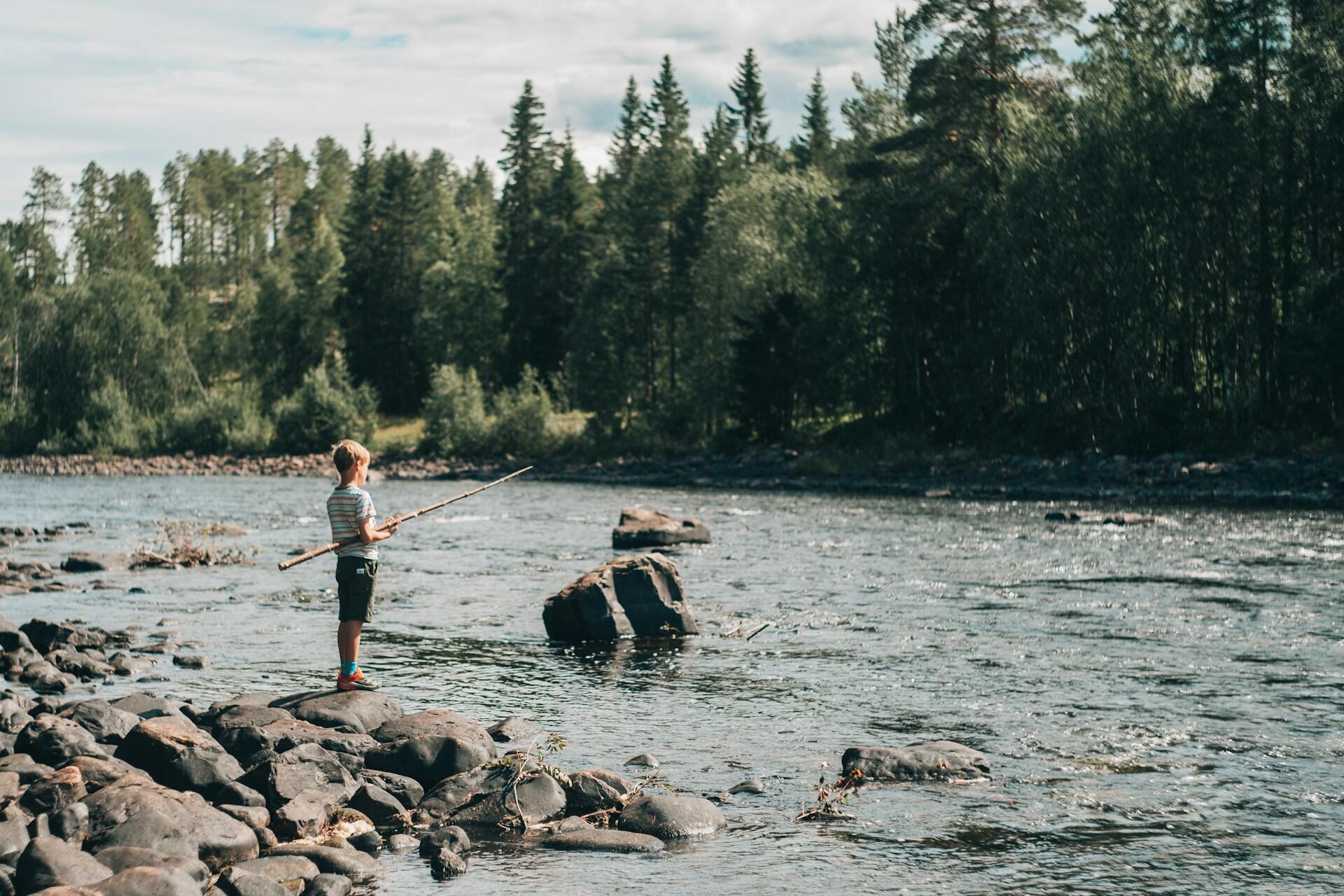 En ung pojke står på en sten vid en flod, med en pinne i handen, omgiven av träd och ett fridfullt naturlandskap.