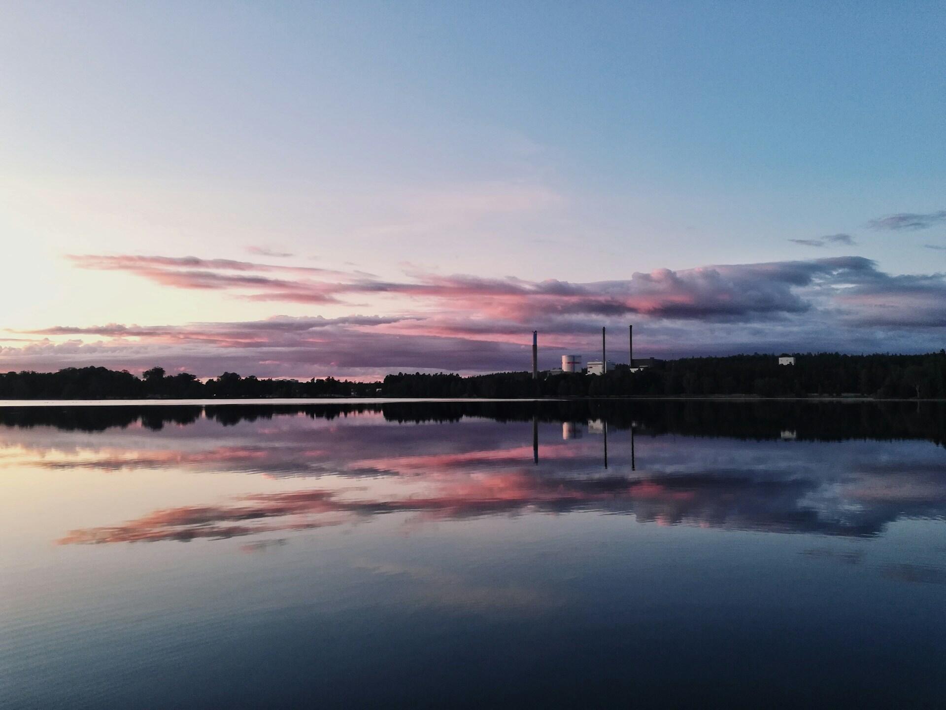 Solnedgång över en lugn sjö, med livfulla rosa och lila moln som speglas i vattnet. Industriella byggnader står i bakgrunden.