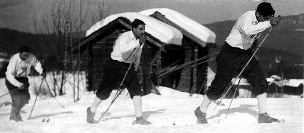 Tre skidåkare tar sig fram i ett snötäckt landskap, med en trästuga i bakgrunden och berg i fjärran. Bilden är från 1948.