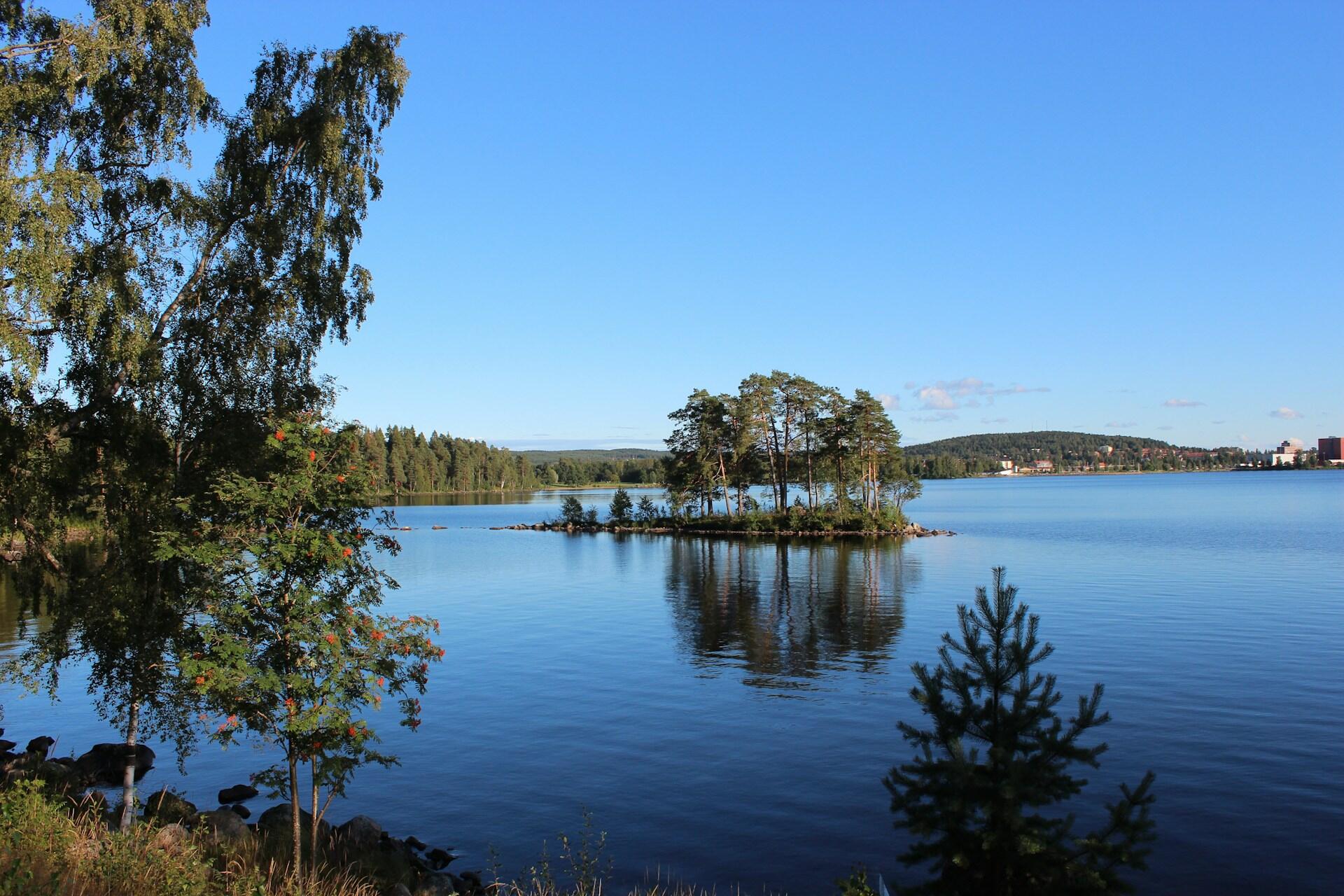 En fridfull sjö med en liten ö med träd, omgiven av lugnt vatten och en klarblå himmel, med skogsklädda kullar i bakgrunden.