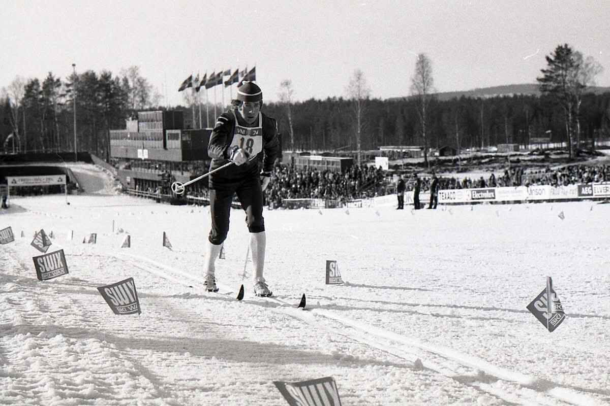 En skidåkare tävlar på en snötäckt bana, omgiven av flaggor och en publik i bakgrunden.