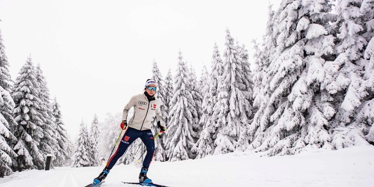 En skidåkare i vinterkläder glider genom ett snötäckt landskap, omgiven av höga, frostiga tallar under en molnig himmel.