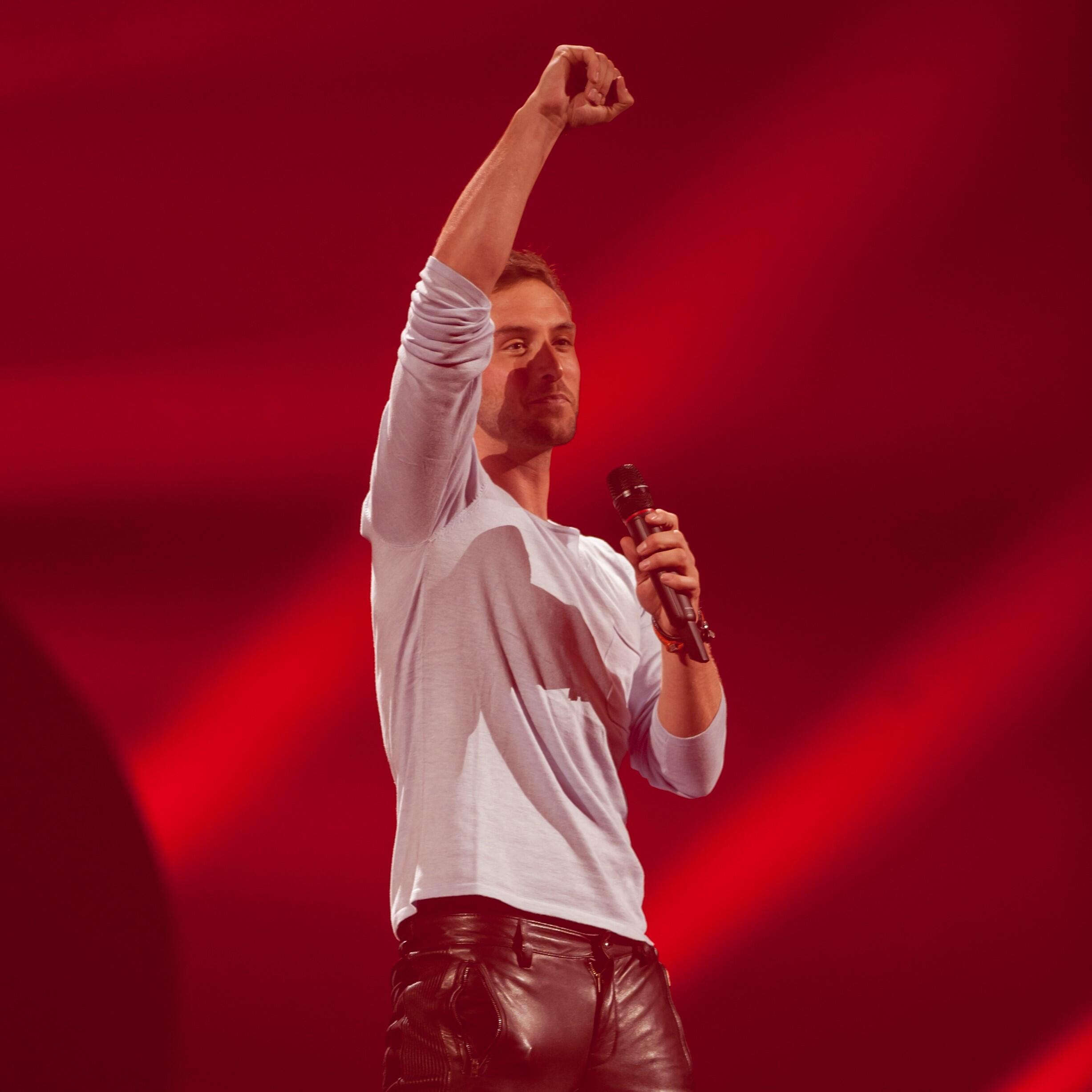Måns Zelmerlöw framför en låt på scen med röd bakgrund.