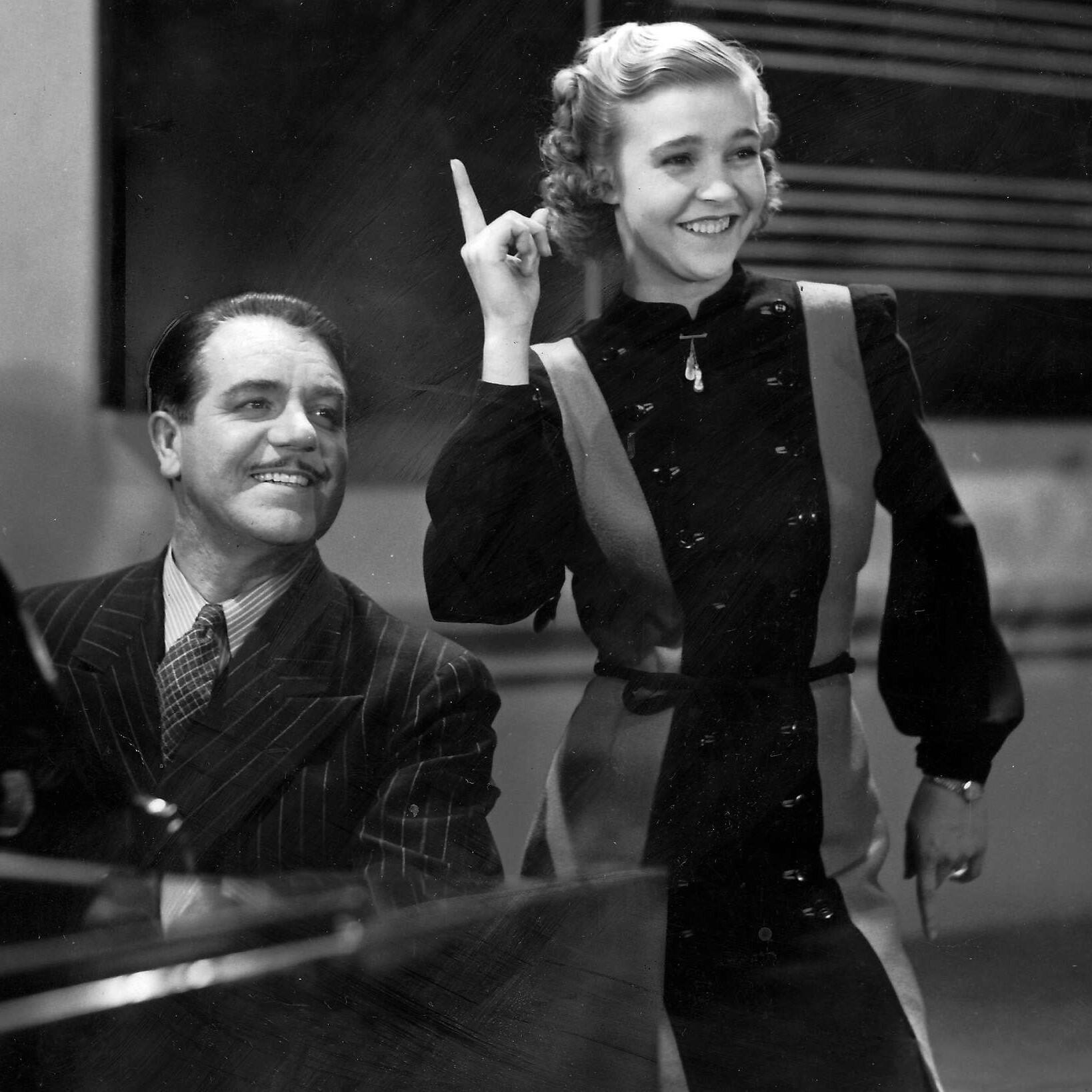 Alice Babs gestikulerar livligt, medan en man sitter bredvid henne, klädd i en randig kostym mot en studiomiljö.