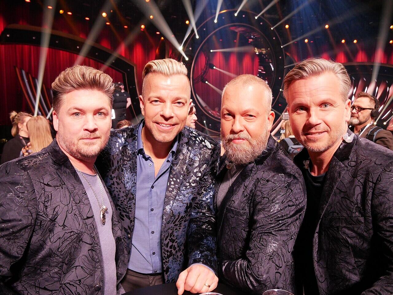 Grupp med fyra män i snygga jackor som poserar tillsammans vid ett livligt evenemang med dramatisk belysning och festlig stämning.